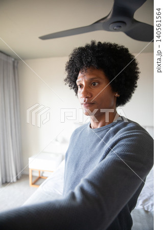 African american man standing beside bed in gray sweater with nightstand, lamp in bedroom 140561564
