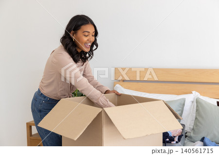 African american woman next to wooden bed unpacking cardboard box in bedroom with suitcase 140561715