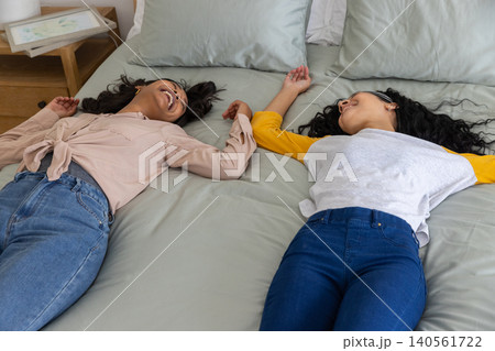 Diverse mom and teenage daughter lying on bed touching hands above heads near table lamp 140561722