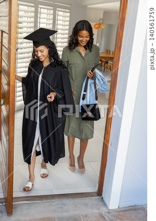 Diverse mom and teenage daughter stepping through doorway into entry with graduation cap and gown 140561759