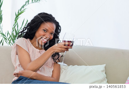 African American woman in 20s sitting on beige sofa wearing pink blouse holding stemmed wine glass African American woman in 20s sitting on beige sofa wearing pink blouse holding stemmed wine glass 140561853