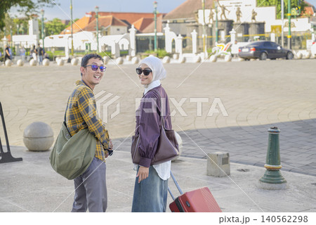A happy young Indonesian southeast asian muslim couple walks through a sunny city plaza with suitcases, smiling at the camera during their vacation travel. Travel or vacation concept A happy young Indonesian southeast asian muslim couple walks through a sunny city plaza with suitcases, smiling at the camera during their vacation travel. Travel or vacation concept 140562298