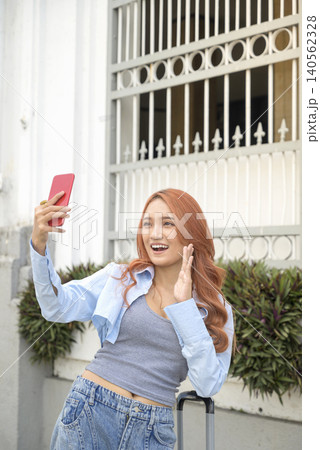 A cheerful young Indonesian southeast asian woman with ginger hair smiles and waves while holding her smartphone for a video call or selfie during her travels. Travel or vacation concept A cheerful young Indonesian southeast asian woman with ginger hair smiles and waves while holding her smartphone for a video call or selfie during her travels. Travel or vacation concept 140562328