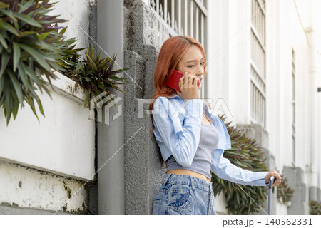 A young Indonesian southeast asian woman with red hair talks on her smartphone while holding a suitcase on a city street during her travels. Travel or vacation concept A young Indonesian southeast asian woman with red hair talks on her smartphone while holding a suitcase on a city street during her travels. Travel or vacation concept 140562331