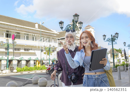 Two happy Indonesian southeast asian female friends, one wearing a hijab, sightsee in a historic city plaza, pointing ahead while holding a digital tablet. Travel or vacation concept Two happy Indonesian southeast asian female friends, one wearing a hijab, sightsee in a historic city plaza, pointing ahead while holding a digital tablet. Travel or vacation concept 140562351