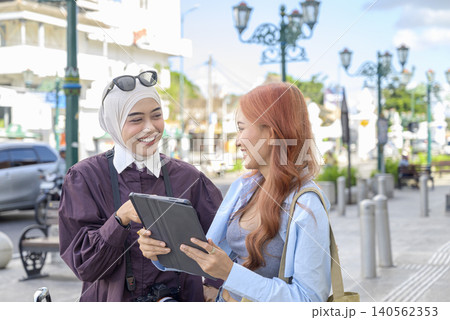 A smiling Indonesian southeast asian muslim woman in a hijab and her friend with colored hair look at a tablet while exploring a city street together. Travel or vacation concept A smiling Indonesian southeast asian muslim woman in a hijab and her friend with colored hair look at a tablet while exploring a city street together. Travel or vacation concept 140562353