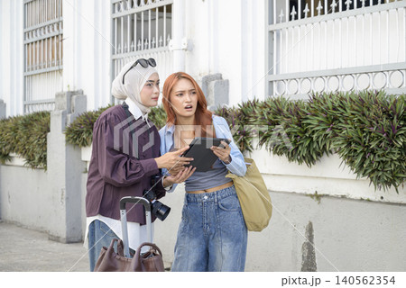 Two female Indonesian southeast asian friends, one wearing a hijab, looking confused while navigating city streets using a digital tablet during their travels. Travel or vacation concept Two female Indonesian southeast asian friends, one wearing a hijab, looking confused while navigating city streets using a digital tablet during their travels. Travel or vacation concept 140562354