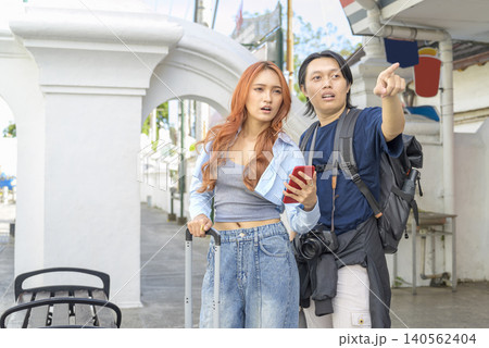 A young Indonesian southeast asian couple traveling together, looking at a smartphone map and pointing directions on an urban street with their luggage. Travel or vacation concept 140562404