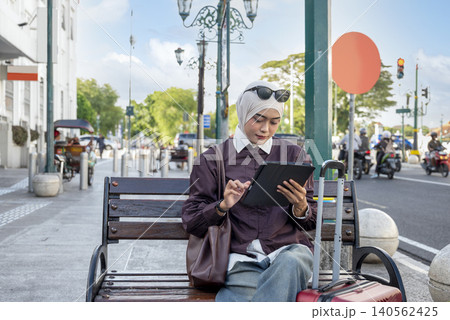 A young Indonesian southeast asian female muslim traveler in a hijab sits on a city bench, using a tablet computer next to her suitcase. Travel or vacation concept 140562425