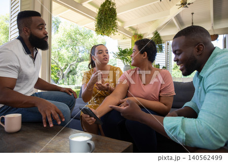 Diverse friends on sofa under porch and holding smartphone by coffee table with ceramic mugs Diverse friends on sofa under porch and holding smartphone by coffee table with ceramic mugs 140562499