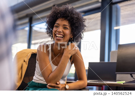African american woman laughing while leaning forward at office desk next to windows with monitors African american woman laughing while leaning forward at office desk next to windows with monitors 140562524