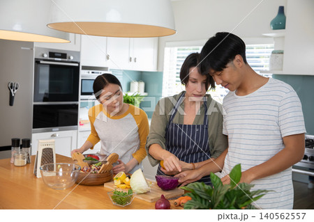 Asian family cooking together at kitchen island chopping vegetables and mixing salad in bowl Asian family cooking together at kitchen island chopping vegetables and mixing salad in bowl 140562557