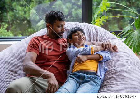 Indian father and son relaxing on beige bean bag at home next to window, potted split-leaf plant Indian father and son relaxing on beige bean bag at home next to window, potted split-leaf plant 140562647