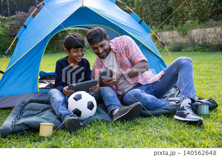 Asian father and son sitting on mat in garden, holding tablet and smartphone next to blue tent Asian father and son sitting on mat in garden, holding tablet and smartphone next to blue tent 140562648
