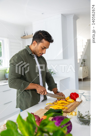 Asian man slicing bell pepper with chef's knife on wooden cutting board at kitchen island Asian man slicing bell pepper with chef's knife on wooden cutting board at kitchen island 140562674