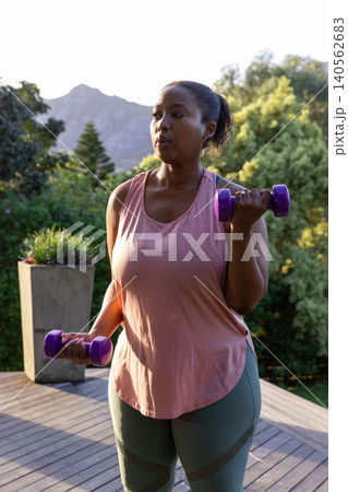African american woman lifting purple dumbbells on backyard deck near planter under mountain sky 140562683