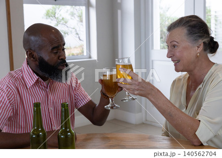 Senior couple clinking beer glasses while sitting at dining table with green bottles Senior couple clinking beer glasses while sitting at dining table with green bottles 140562704