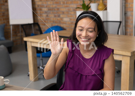 Asian adult woman sitting at office table waving at laptop screen Asian adult woman sitting at office table waving at laptop screen 140562709