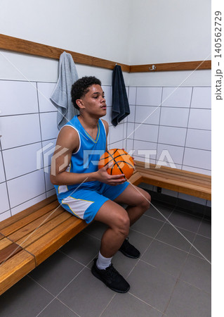 African american teenage male athlete wearing jersey sitting in locker room with basketball 140562729