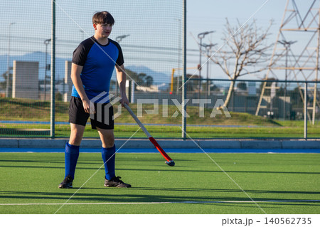 Male field hockey player gripping stick with red taped head and standing on turf pitch Male field hockey player gripping stick with red taped head and standing on turf pitch 140562735