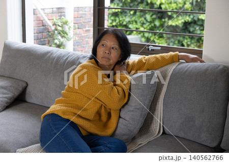 Senior asian woman in mustard sweater, jeans reclining on sofa cushion gazing through blinds 140562766