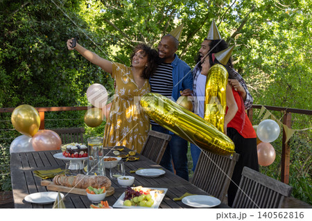 Diverse friends wearing party hats taking selfie on deck with smartphone by table near balloons Diverse friends wearing party hats taking selfie on deck with smartphone by table near balloons 140562816