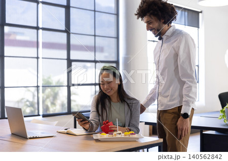 Diverse coworkers leaning in office with laptop and treat tray with smartphone gift box smiling Diverse coworkers leaning in office with laptop and treat tray with smartphone gift box smiling 140562842