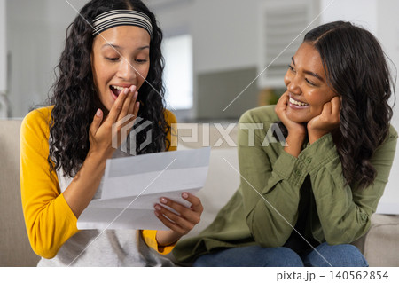 Diverse mom and teenage daughter sitting on couch in living room reading letter smiling 140562854
