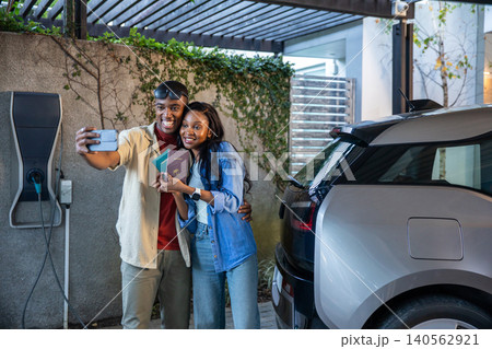 African American couple standing under pergola taking selfie holding passports next to EV charger 140562921