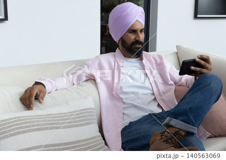 Indian man sitting on sofa in living room holding smartphone, gazing at screen, copy space Indian man sitting on sofa in living room holding smartphone, gazing at screen, copy space 140563069