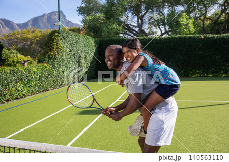 Asian girl riding piggyback on African American father gripping tennis racket by net on court Asian girl riding piggyback on African American father gripping tennis racket by net on court 140563110