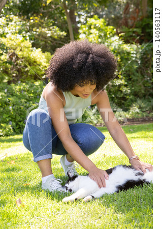 African american woman crouching on green lawn in private garden petting black-and-white cat 140563117