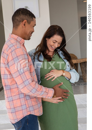Diverse couple standing in home placing palms on pregnant belly by botanical print, table and rug 140563199