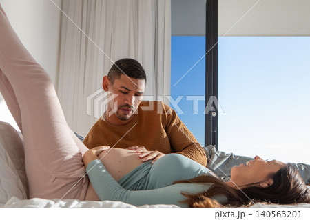 Diverse expecting couple sharing moment reclining on bed in modern bedroom by sliding glass window Diverse expecting couple sharing moment reclining on bed in modern bedroom by sliding glass window 140563201