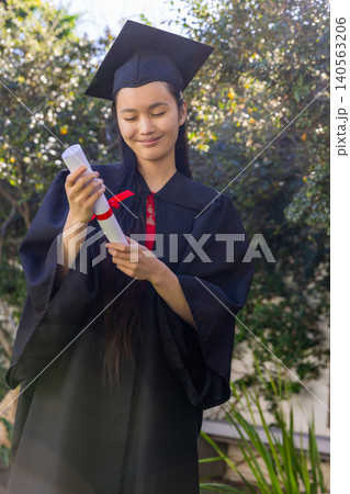 Asian female graduate holding diploma tied with red ribbon wearing black gown and cap Asian female graduate holding diploma tied with red ribbon wearing black gown and cap 140563206