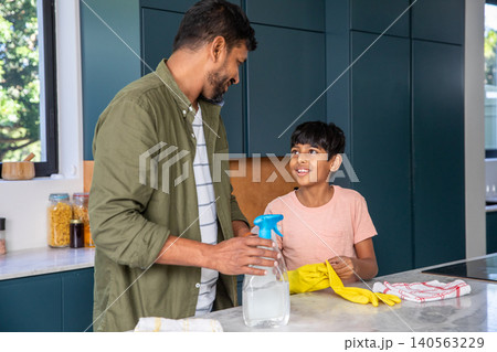 Indian father with son standing at countertop in home kitchen spraying cleaner while wearing gloves 140563229
