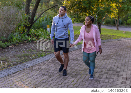 Diverse couple walking hand in hand along path in park among shrubs under sunlit trees 140563268