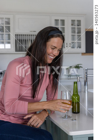Woman in her 30s sitting in kitchen holding beer with green beer bottle on countertop Woman in her 30s sitting in kitchen holding beer with green beer bottle on countertop 140563273
