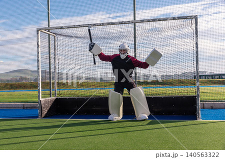 Standing in front of hockey goal, male goalkeeper in protective gear holding stick on turf Standing in front of hockey goal, male goalkeeper in protective gear holding stick on turf 140563322