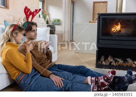 Couple holding ceramic mugs and wearing antler headbands sitting by fireplace in living room Couple holding ceramic mugs and wearing antler headbands sitting by fireplace in living room 140563351