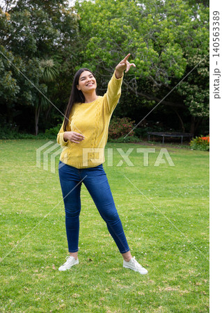 Woman standing on lawn wearing mustard sweater and jeans pointing in garden with trampoline 140563389