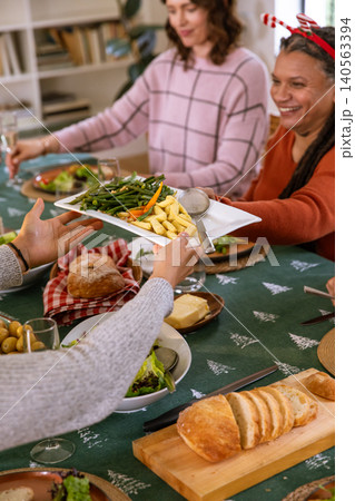 Diverse family in festive sweaters passing platter of green beans, potatoes around dining table 140563394