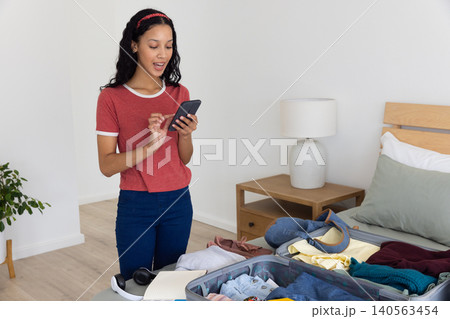 Teenage girl beside open suitcase on bed in bedroom tapping smartphone near headphones 140563454