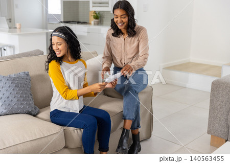 Diverse mom and teenage daughter sitting on sofa in living room exchanging paper, copy space 140563455