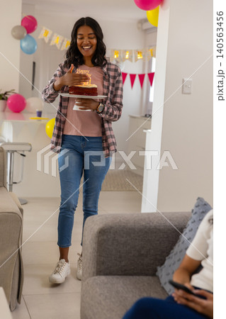 Mother carrying cake with lit candles at home with balloons, banner, one holding smartphone Mother carrying cake with lit candles at home with balloons, banner, one holding smartphone 140563456