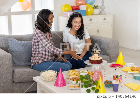 Diverse mom and teenage daughter celebrating birthday on gray sofa in living room with cake 140564140