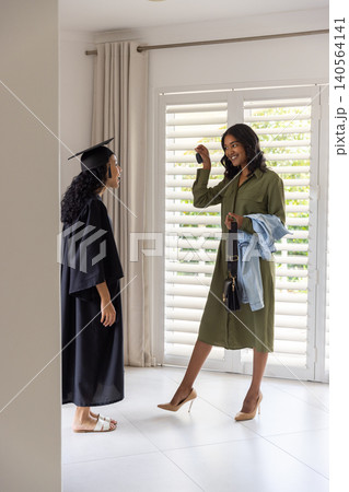 Diverse mom and teenage daughter exchanging keys at entrance, daughter in black cap and gown 140564141