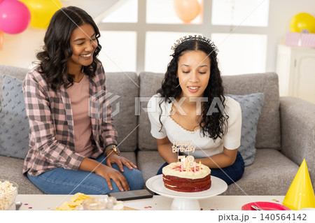 Diverse mom and teenage daughter blowing candles on red velvet birthday cake under balloons Diverse mom and teenage daughter blowing candles on red velvet birthday cake under balloons 140564142