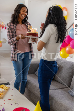 Diverse mom and teenage daughter celebrating birthday in living room with lit cake and balloons 140564143