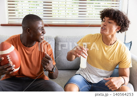 Diverse male friends sitting on couch in living room holding football, raising bottled beverage 140564253